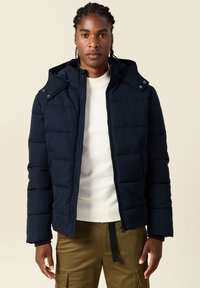 Marinblå puffjacka med huva, vadderad design, lätt tyg, frontdragkedja och tryckknappar. Buren över en vit tröja.