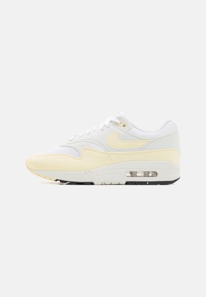 Białe sneakersy z żółtymi akcentami, wykonane z siateczki i zamszu, o zaokrąglonym kształcie, z widoczną jednostką powietrzną w podeszwie oraz kontrastującą czarną podeszwą.