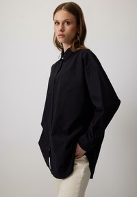Donna con capelli lunghi, che indossa una camicia nera oversize con bottoni e pantaloni beige, in piedi di lato con una mano in tasca su uno sfondo semplice.