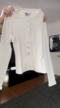 Valge pika varrukaga Henley särk seitsme nupuga, mida hoiab kahe käega elutoa keskkonnas, kus on puidust põrand ja raamitud seinamaalid.