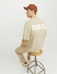 Beige cotton t-shirt with white text on the back, beige pants, and multicolored sneakers. Brown cap, seated on a wooden stool.