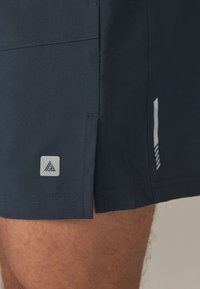Pantaloncini sportivi blu navy scuro con una texture liscia, caratterizzati da una piccola toppa con il logo e un dettaglio a righe bianche sul lato.