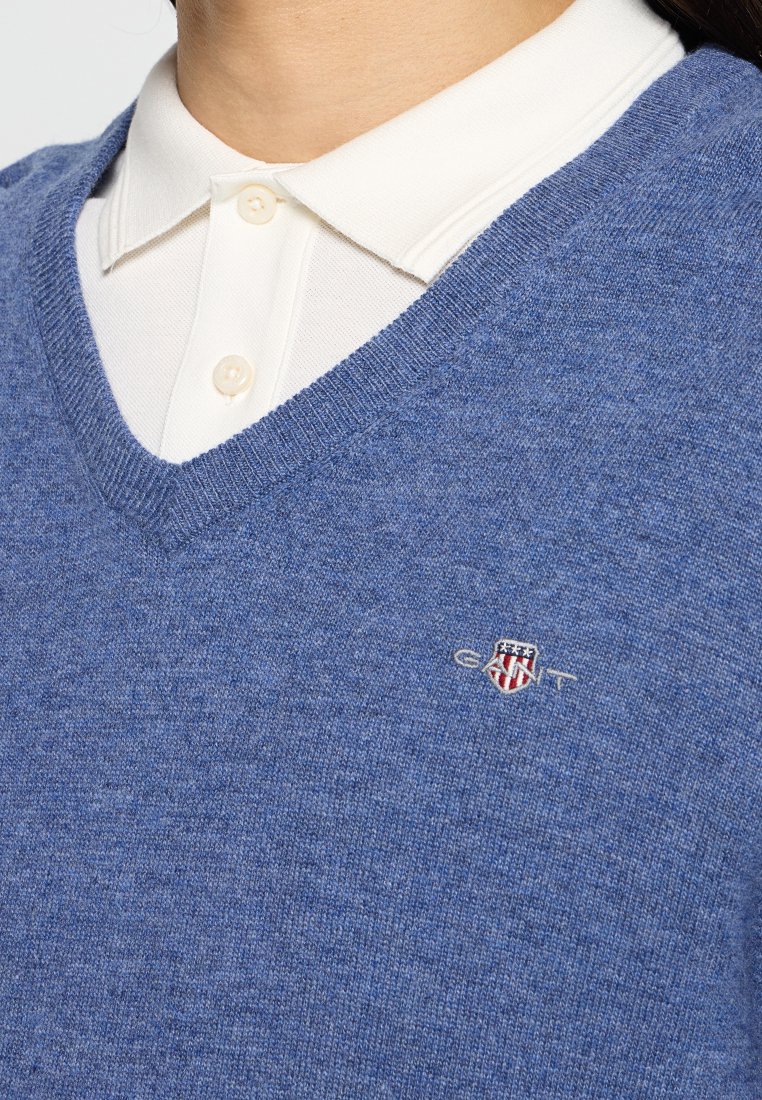 Niebieski sweter dzianinowy z dekoltem w serek, z białą koszulą z kołnierzykiem pod spodem oraz haftowanym logo na lewej piersi.