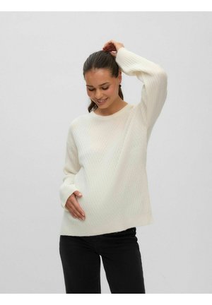 Femme souriante en pull en maille blanc et pantalon noir touche son ventre d'une main tandis que l'autre main soulève ses cheveux.
