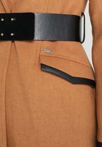 Manteau en laine couleur camel avec une large ceinture en cuir noir et un rabat de poche assorti bordé de noir, avec une petite plaque métallique « Guess ».