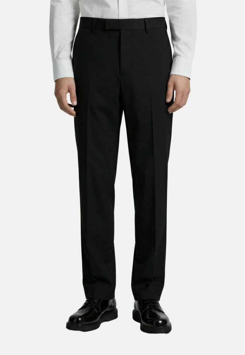 Hombre que lleva pantalones formales negros, zapatos de vestir de cuero negros y una camisa blanca de manga larga, de pie frente a un fondo liso.