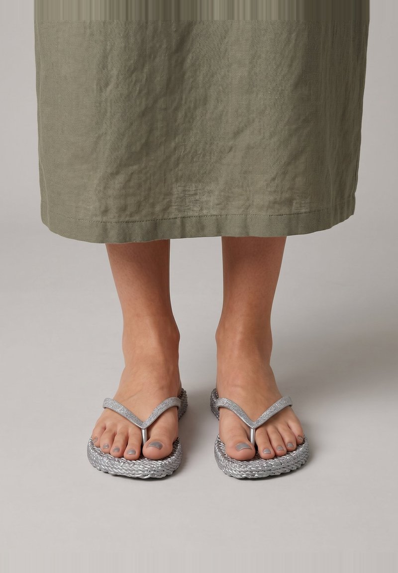 Voeten met zilverkleurige gevlochten teenslippers met grijze nagellak, onder een lange olijfgroene rok, staand op een eenvoudige lichte ondergrond.