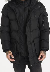 Svart puffjacka med dragkedja och knappstängning, med en foderhuva av päls, flera fickor och en logotyp på ärmen. Slät yta.