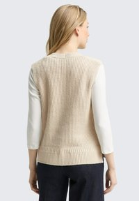 Beige gebreide mouwloze trui met getextureerde ribbels en een smalle tailleband, gedragen over een witte langekraag shirt met een ronde halslijn.