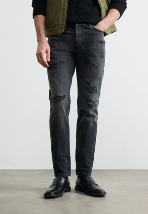 Mann trägt schwarze zerrissene Jeans, schwarze Schuhe, schwarzen Pullover und olivgrüne Weste, steht mit einer Hand in der Tasche.