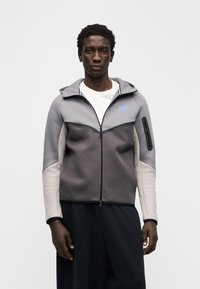 Pelēka sporta jaka ar rāvējslēdzēju un kapuci, kas ir ar kontrastējošiem paneļiem, gaiši pelēkām piedurknēm un mazu zilu logotipu uz krūtīm.