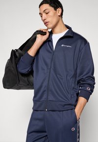 Μπλε ναυτικό τζάκετ με φερμουάρ και λευκό λογότυπο, παντελόνια που ταιριάζουν και ρίγες στις πλευρές. Το μοντέλο κρατά μια μαύρη τσάντα duffel με πάνελ από δίχτυ. Ελαφρύ ύφασμα.