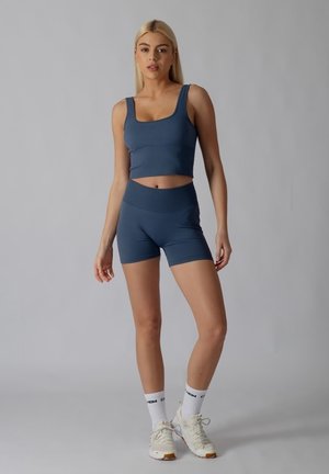 Młoda kobieta stoi w szarym sportowym crop topie i szortach, w białych skarpetkach i butach sportowych na jednolitym szarym tle.