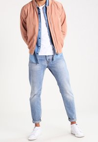 Jasnoróżowa kurtka bomberka na koszuli dżinsowej, biała koszulka, jasnoniebieskie dżinsy i białe trampki. Gładkie tekstury i klasyczne dopasowanie.