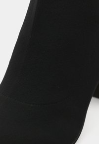 Gros plan sur une botte cheville en tissu noir avec talon bloc, montrant une texture lisse et un détail de couture incurvée près de la base.