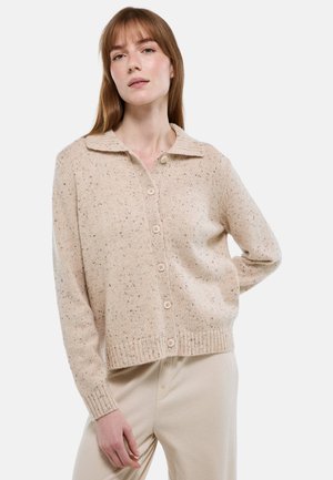 Femme aux cheveux bruns clairs et raides portant un cardigan beige à boutons tachetés et un pantalon assorti sur fond blanc, debout avec une main derrière.