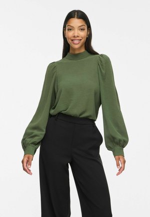 Vrouw glimlachend, draagt een groene blouse met lange mouwen, pofmouwen en een hoge hals, gecombineerd met zwarte hoge taillebroeken.