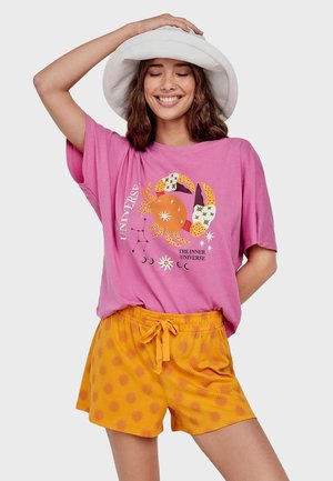 Giovane donna che sorride con gli occhi chiusi, indossa un cappello bianco, una t-shirt rosa con grafica di granchio e pantaloncini arancioni con motivo sole.
