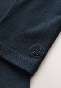 Tamsiai mėlynas medvilninis audinys iš arti, su tekstūruotu įsiuviniu logotipu šviesesnės spalvos ir matomomis siūlėmis palei kraštą.