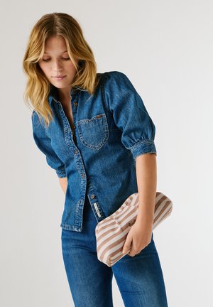 Jeune femme en chemise en jean et jean tenant une pochette beige à rayures blanches sur un fond uni.