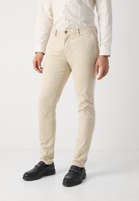 Chinos en coton beige avec une coupe slim, dotés de poches latérales et d'une fermeture à bouton à l'avant, accompagnés de mocassins en cuir noir.