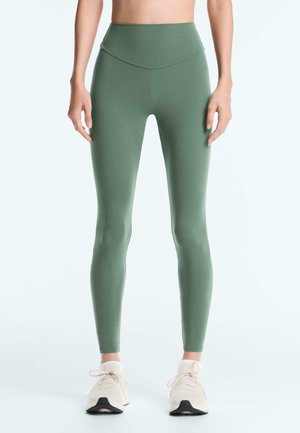 Osoba w oliwkowozielonych legginsach z wysokim stanem i białych butach sportowych, stojąca z luźno opadającymi ramionami.