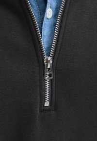 Lähivaade osaliselt lukustamata hõbedasest lukkudest mustal kangal, mis paljastab sinise nööbitud särgi allpool.