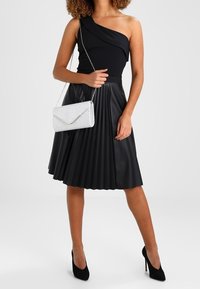 Femme portant un haut noir à une épaule, une jupe plissée noire, des talons hauts noirs et tenant une pochette à paillettes argentées avec une chaîne.