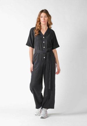 Woman with wavy hair wearing a black button-up jumpsuit with a waist tie and white sneakers, standing on a plain light background.