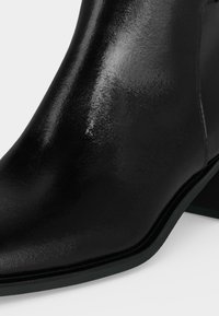 Gros plan d'une bottine en cuir noir brillant avec un bout arrondi et un petit talon bloc sur fond blanc.