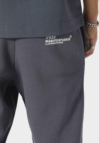 Pantalons de jogging en coton gris foncé avec un logo en texte blanc "IRASUTOSTUDIOS" et des caractères japonais sur la poche arrière. Texture lisse, coupe décontractée.