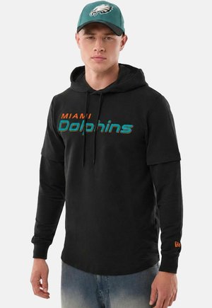 Giovane uomo con una felpa nera dei Miami Dolphins con maniche a strati e un cappellino verde dei Philadelphia Eagles, in piedi su uno sfondo semplice.
