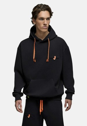 Jeune homme portant un sweat à capuche noir et un pantalon avec des cordons orange vif et de petits détails de poche, debout devant un fond uni.