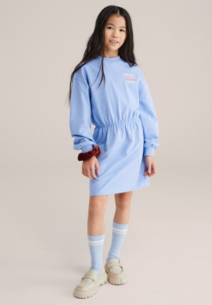 Zils kokvilnas kleita ar garām piedurknēm, elastīgu vidukli un tekstu uz krūtīm. Kombinēta ar ceļgalu augstām svītrainām zeķēm un masīvām kurpēm.