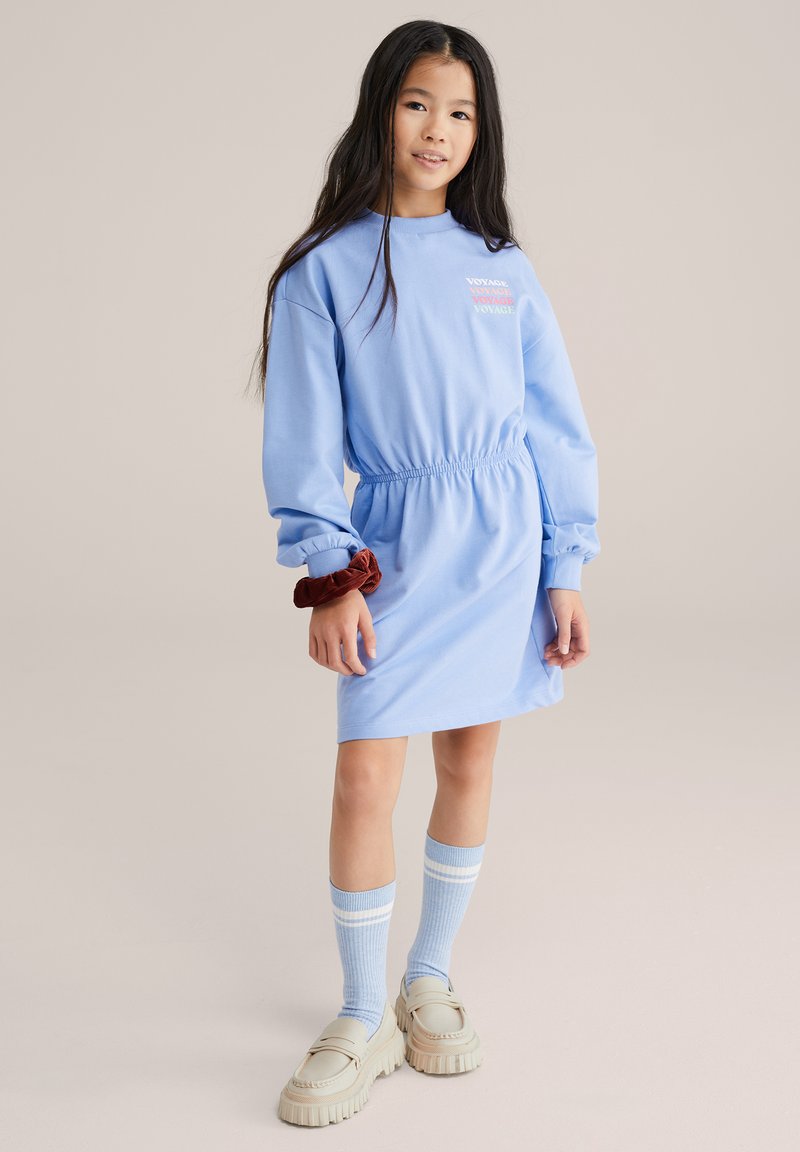 Vestido azul de algodón con mangas largas, cintura elástica y detalle de texto en el pecho. Combinado con calcetas a rayas hasta la rodilla y zapatos gruesos.