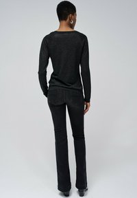 Person mit kurzen Haaren, die silberne Creolen, ein schwarzes Langarmshirt und schwarze enge Jeans trägt und mit dem Rücken zur Kamera steht.