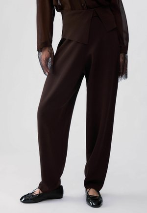 Person trägt dunkelbraune, weit geschnittene Hose und durchsichtiges Spitzen-Longsleeve mit schwarzen Ballerinas vor hellem Hintergrund.
