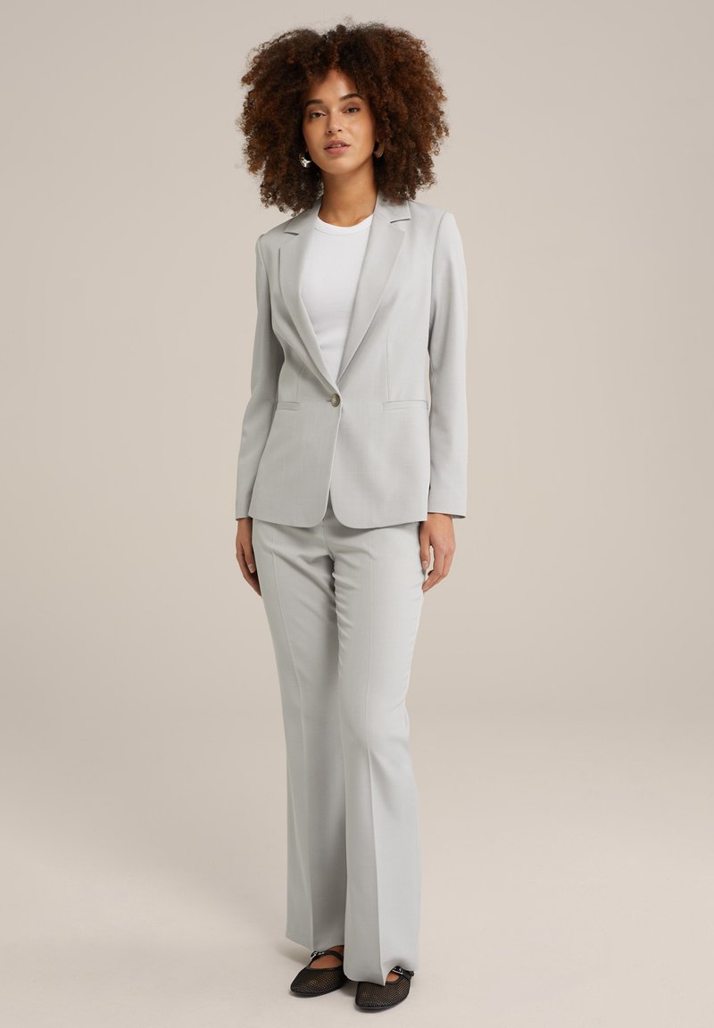 Costume gris clair avec une jacket ajustée et un pantalon évasé. Le matériel semble lisse, avec des coutures subtiles et un détail de bouton unique. ballerines noires.