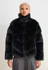 Crni puffer kaput od umjetnog krzna s visokim ovratnikom, s kućastim dizajnom, zatvaranjem na zip i mekšim teksturom, nosi se preko sive suknje.