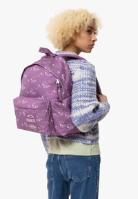 Mochila morada con patrones gráficos blancos, que cuenta con correas ajustables, un bolsillo frontal y una tela texturizada. Logo visible en la parte frontal.