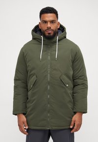 Grønne polstrede parka-jakke med hætte, lynlås lukning, to forlommer og snore. Glat tekstur med grå foer.