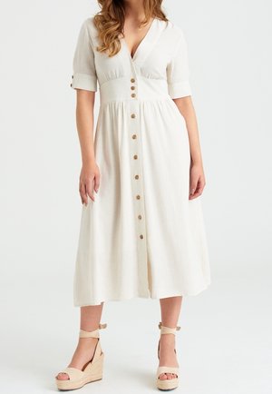 Vrouw in een witte midi-jurk met houten knopen aan de voorkant en beige sleehakken met enkelbandjes tegen een effen witte achtergrond.