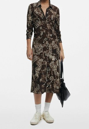 Kvinde iført en brun mønstret midi-skjortekjole med lange ærmer, hvide sokker, beige sko og holder en sort håndtaske.