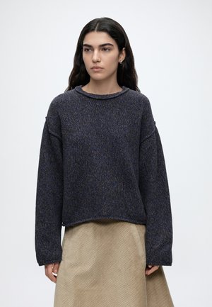 Jeune femme aux longs cheveux foncés portant un pull en maille bleu foncé ample et une jupe en velours côtelé beige, debout devant un fond uni.