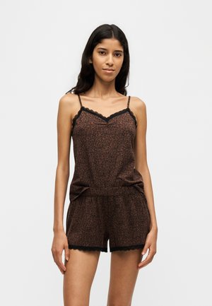 Jonge vrouw met lang donker haar, gekleed in een bruine leopardprint camisole en bijpassende korte broek met zwarte kanten rand, staand tegen een effen achtergrond.