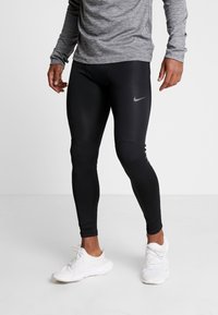 Persona che indossa leggings atletici neri Nike e sneakers bianche, in piedi davanti a uno sfondo grigio uniforme.