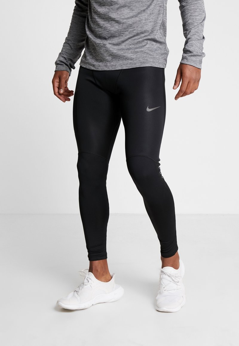 Persona che indossa leggings atletici neri Nike e sneakers bianche, in piedi davanti a uno sfondo grigio uniforme.