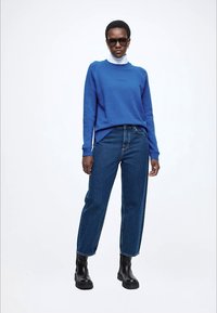 Camisola azul, top branco de gola alta, jeans de ganga e botins pretos até ao tornozelo. Características incluem mangas compridas e um corte descontraído.