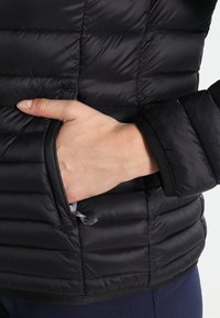 Zwarte pufferjack met horizontale naden, een zijzak en elastische manchetten. Het materiaal lijkt glad met een lichtgewicht textuur.