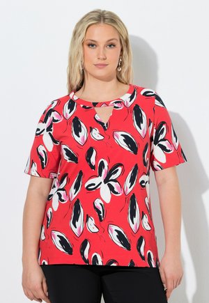 Donna bionda con una maglietta rossa a maniche corte con motivi floreali astratti in bianco e nero e una piccola apertura a forma di goccia sul collo.